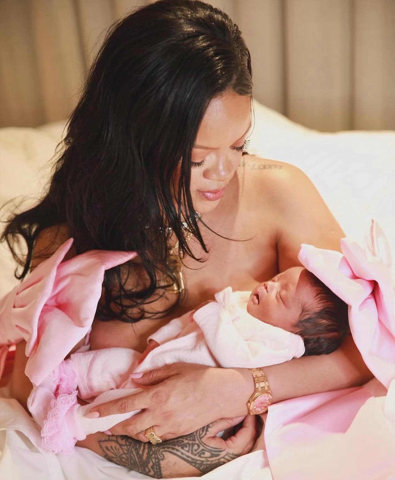 Rihanna holding her new born baby wrapped in pink, smiling lovingly while sitting on a bed. The Singer wears a pink outfit and gold jewelry, celebrating motherhood in a tender moment
