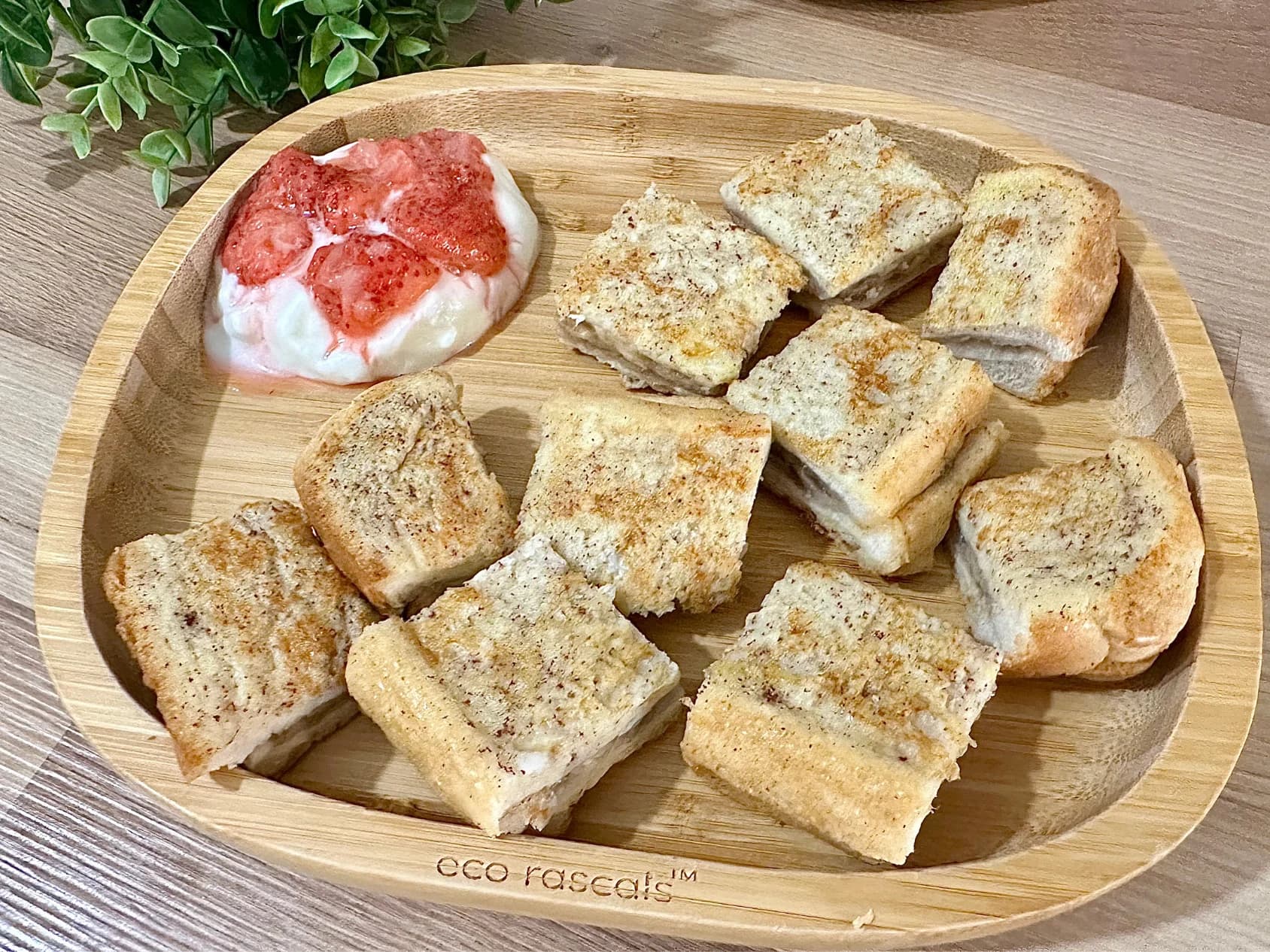 Overhead view of a wooden "eco rascals" plate with neatly arranged squares of banana French toast and a side of yogurt topped with mashed strawberries, ideal for baby-led weaning.