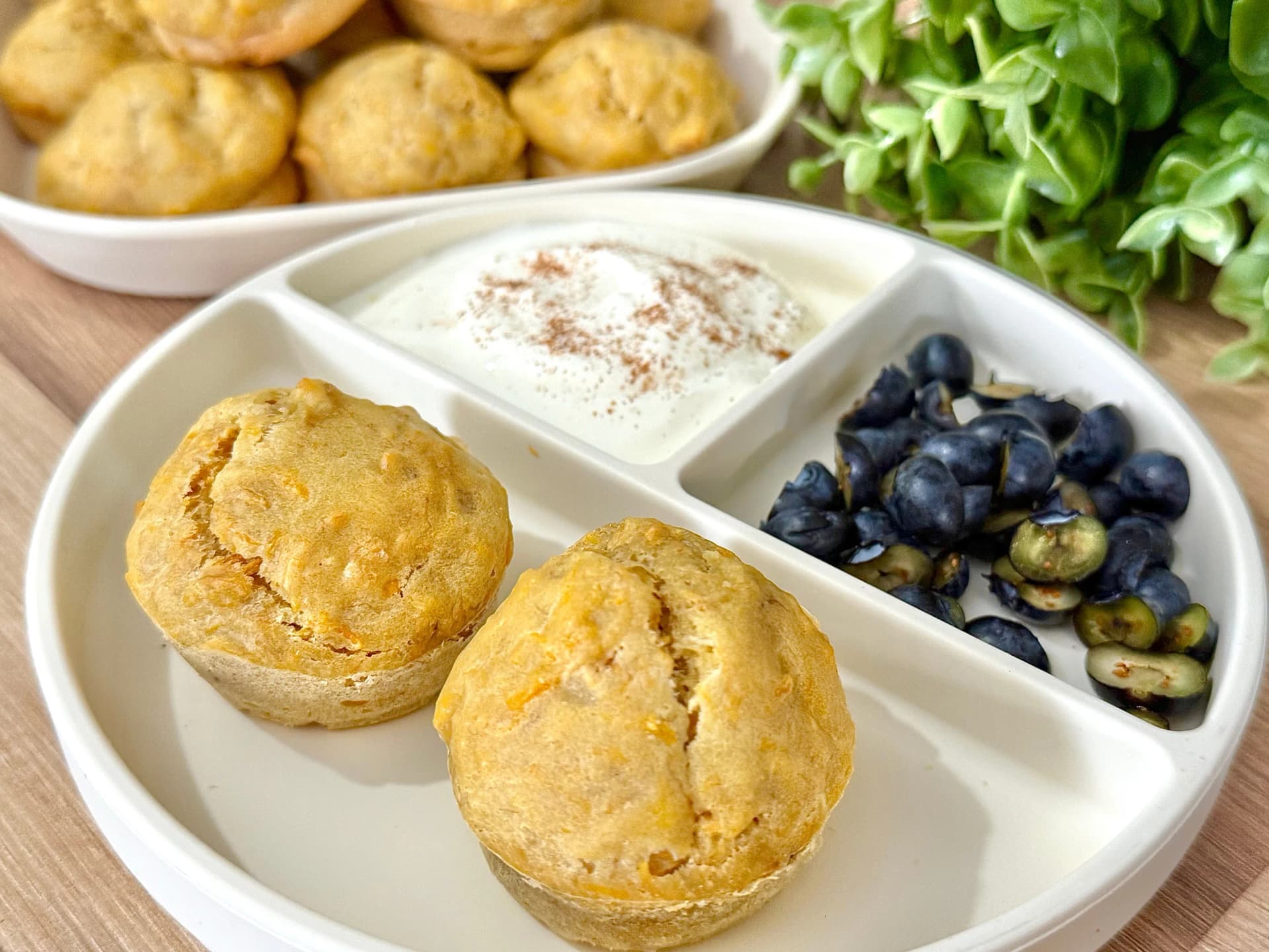 "Delicious golden banana muffins on a white plate, accompanied by fresh blueberries and a dollop of yogurt sprinkled with cinnamon. Ideal for breakfast or a sweet treat."