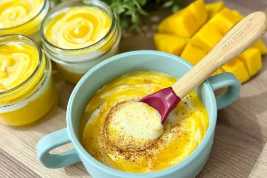 Close-up of homemade mango yogurt in a baby-friendly blue bowl with an Eco Rascals bamboo spoon, swirled with cinnamon, and individual servings in jars with fresh mango cubes in the background. Perfect for baby-led weaning or a healthy snack.