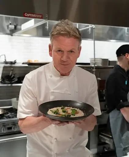 Celebrity chef Gordon Ramsay standing confidently in a professional kitchen, wearing his signature white chef’s jacket, surrounded by cooking equipment and ingredients.