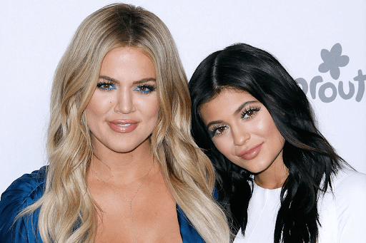 Close-up photograph of sisters Khloe Kardashian, with long blonde hair and blue eyeliner, standing next to Kylie Jenner, who has long black hair and is wearing white.