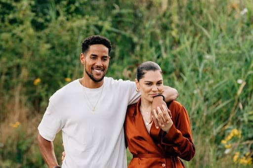 Jessie J walks outdoors with her partner, who has his arm around her shoulder, sharing a warm and relaxed moment in a lush green setting.