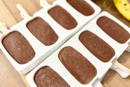 "Close-up of two white silicone popsicle molds filled with frozen, creamy chocolate mixture, with wooden sticks inserted. The surface shows a light frosty layer, indicating they are ready to be fully frozen or just removed from the freezer. Perfect for homemade chocolate ice pops."