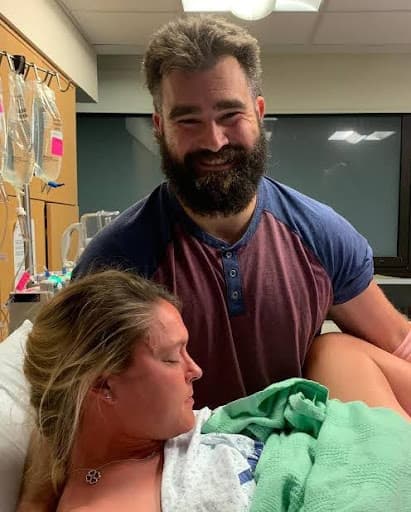 "Expecting parents in a hospital room during labor – father standing behind, smiling and supporting mother lying in bed with a green blanket, captured in a joyful and intimate moment before childbirth."