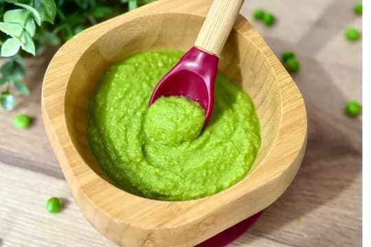 Close-up of smooth, vibrant green pea puree served in a wooden baby bowl with a red and bamboo spoon. Loose peas are scattered on the wooden surface, showcasing this healthy homemade first food for infants and toddlers.