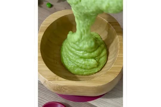 "Smooth green pea and potato puree being poured into a wooden baby bowl, illustrating homemade baby food or a creamy vegetable mash."