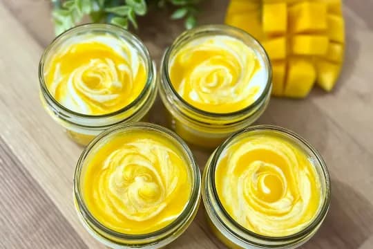 Four glass jars of homemade mango yogurt with beautiful yellow and white swirls, garnished with a mango cube in the background, ready to serve as a dessert or snack.
