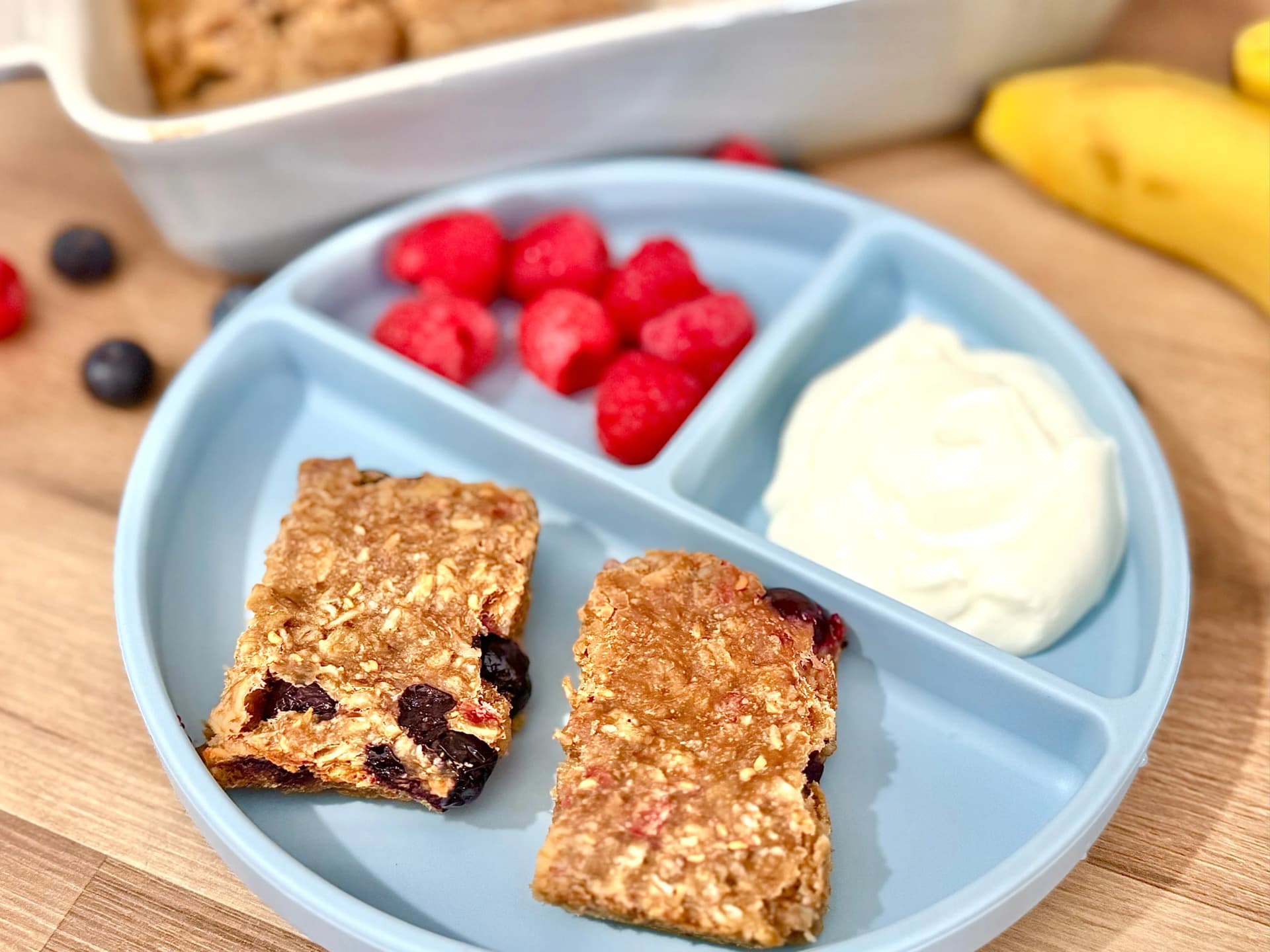 Healthy breakfast plate with baked protein berry oat bars, fresh raspberries, Greek yogurt, and banana, served on a blue divided plate.