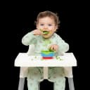 Happy, messy toddler in a high chair feeding themselves peas with a spoon, illustrating the self-feeding milestone and developing fine motor skills.