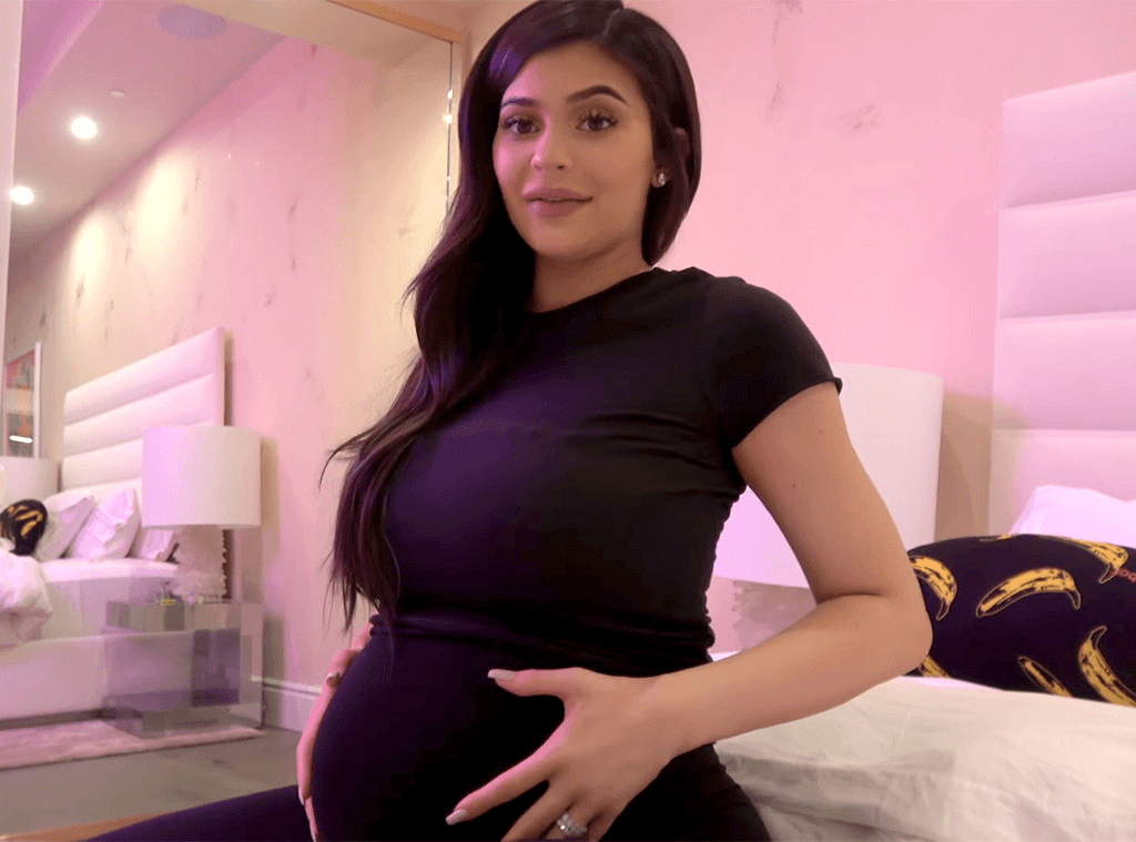 Kylie Jenner sitting in a pink-lit bedroom, wearing a black t-shirt and holding her visibly pregnant belly with both hands.