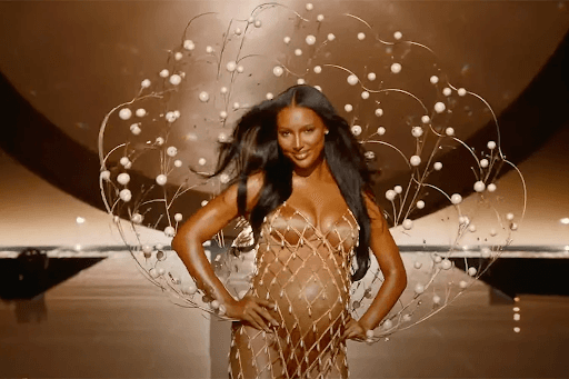 A pregnant woman confidently walking on a runway wearing a gold mesh dress with an artistic circular design featuring pearl-like ornaments surrounding her.