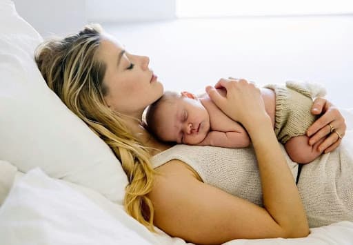A woman lying on a bed, peacefully holding a sleeping baby on her chest, both resting in a serene and intimate moment.