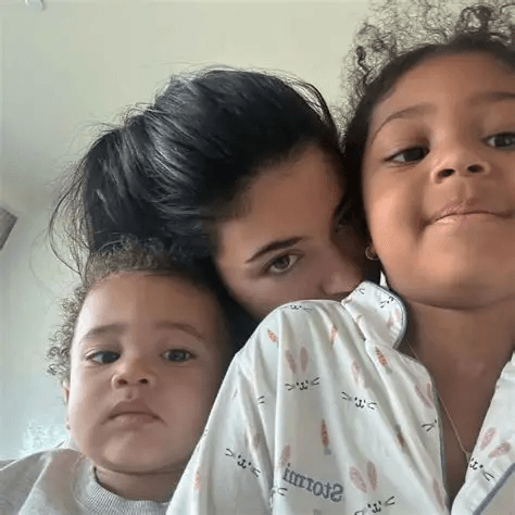 Close-up selfie of Kylie Jenner with her two young children, Stormi Webster and Aire Webster. The three are cuddled together, with Kylie's face peeking over the children's shoulders.