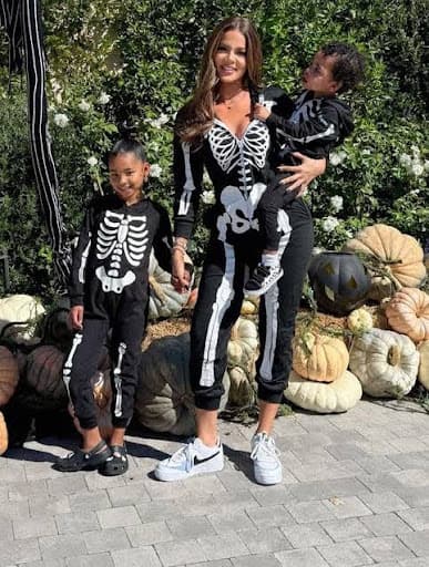 Woman and two children in matching skeleton costumes for Halloween, posing outdoors with pumpkins