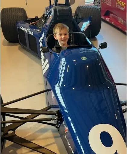 A young boy smiling while sitting in the driver’s seat of a blue Formula-style race car, hands on the steering wheel, surrounded by other race cars in a brightly lit garage.