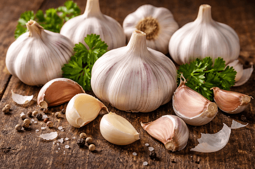 Whole raw garlic bulbs and peeled cloves on a rustic wooden table with fresh parsley sprigs and peppercorns.