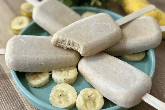 Finished banana popsicles being removed from molds, with one held up and ready to eat, and warm water running over a mold to help release it.