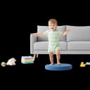 Flat illustration of a joyful baby or toddler with light skin, wearing a blue shirt and white diaper, standing and trying to balance with arms outstretched. This image represents a key gross motor skill milestone, like walking or standing unsupported.