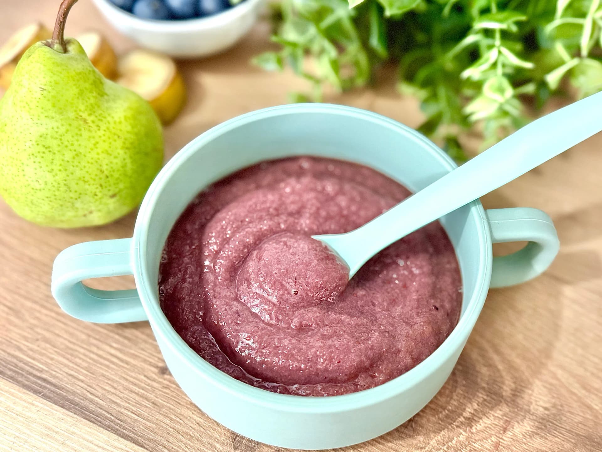 A bowl of Berry Pear Puree for babies, sitting on a wooden table with a pear and blueberries nearby.