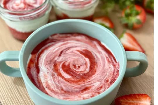 Close-up overhead shot of creamy strawberry yogurt swirled with red fruit puree in a light blue baby bowl. In the background, two layered strawberry yogurt parfaits in glass jars and fresh sliced strawberries are visible on a wooden surface. Ideal for a healthy breakfast, snack, or baby food."