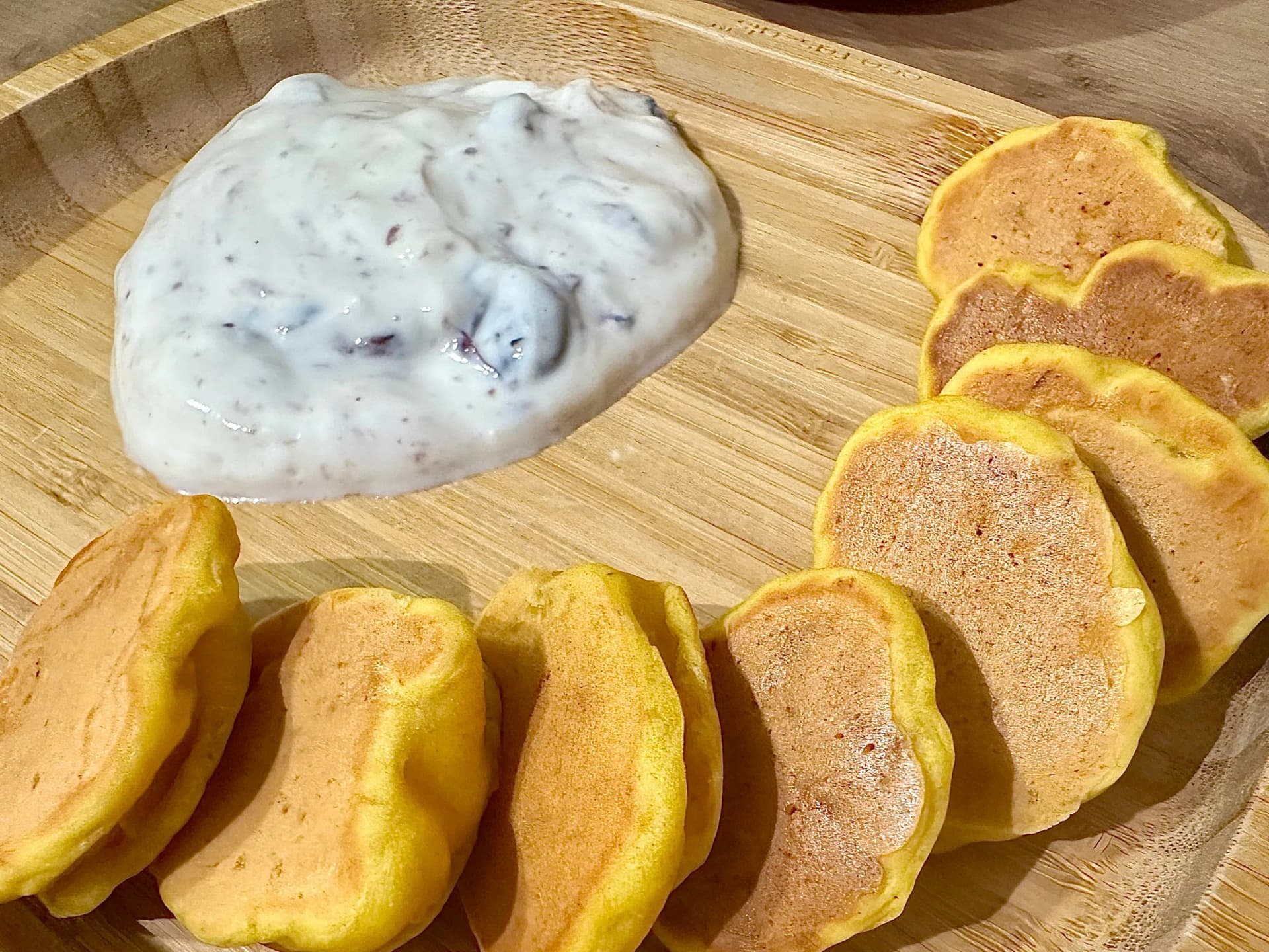Overhead view of a wooden serving tray with a row of golden mini pumpkin pancakes and a dollop of white, berry-swirled yogurt.