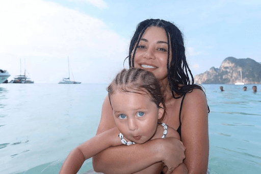 Martha Kalifatidis, smiling and holding her young child in clear blue water during a beach outing, as she and Michael Brunelli announce they are expecting their second baby.