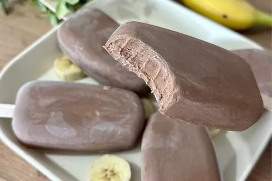 "Close-up of a creamy chocolate popsicle with a bite taken out, showing its smooth, rich texture. Several other chocolate popsicles and sliced bananas are visible on a white rectangular plate in the background. Perfect for a refreshing homemade dessert or healthy summer treat."