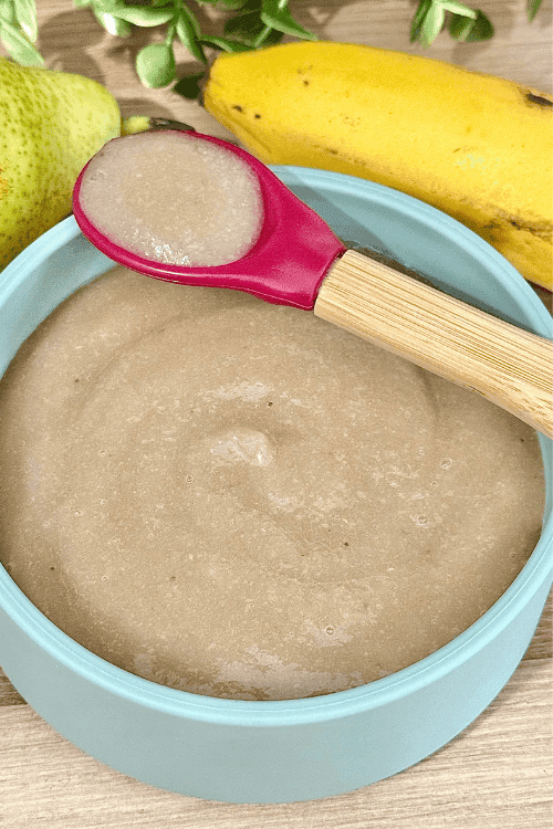 Bowl of smooth pear and banana purée in a light blue handled bowl with a bamboo spoon, placed on a wooden surface with fresh pear and banana slices in the background.