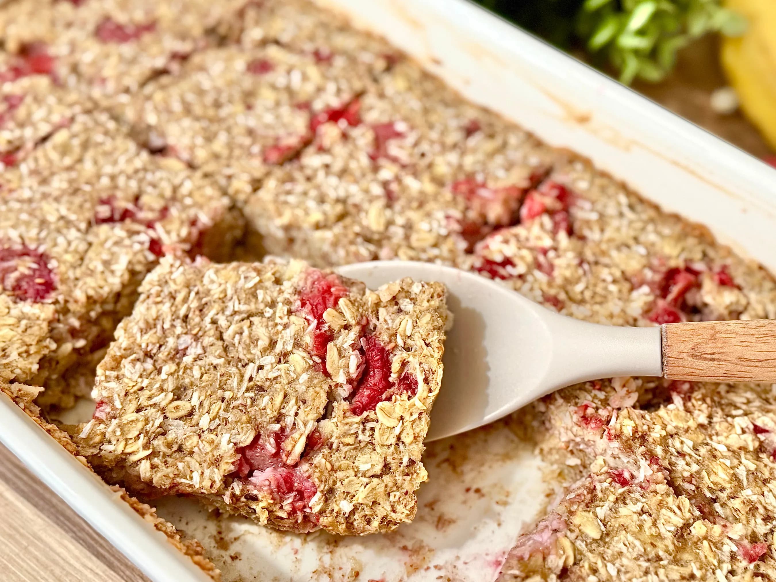 Homemade coconut and raspberry oat bars in a baking dish, with one square lifted on a spatula, showing a chewy texture and fresh fruit pieces.