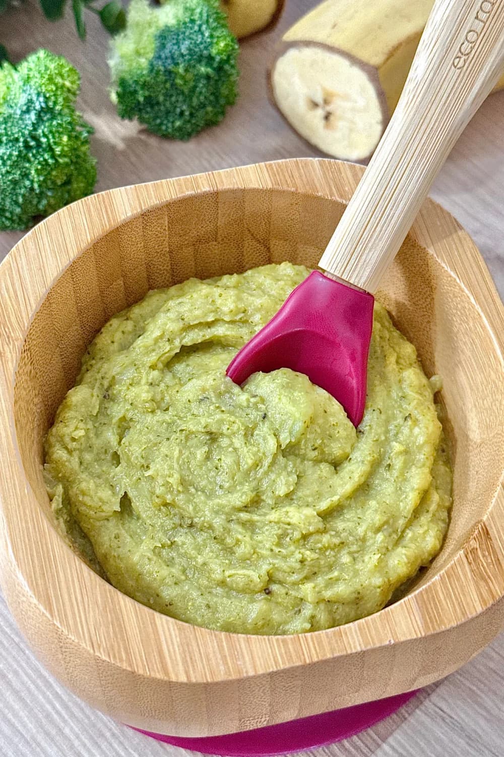 A close-up of a textured green purée in a wooden bowl with a pink baby spoon. The background shows raw plantain and broccoli florets.