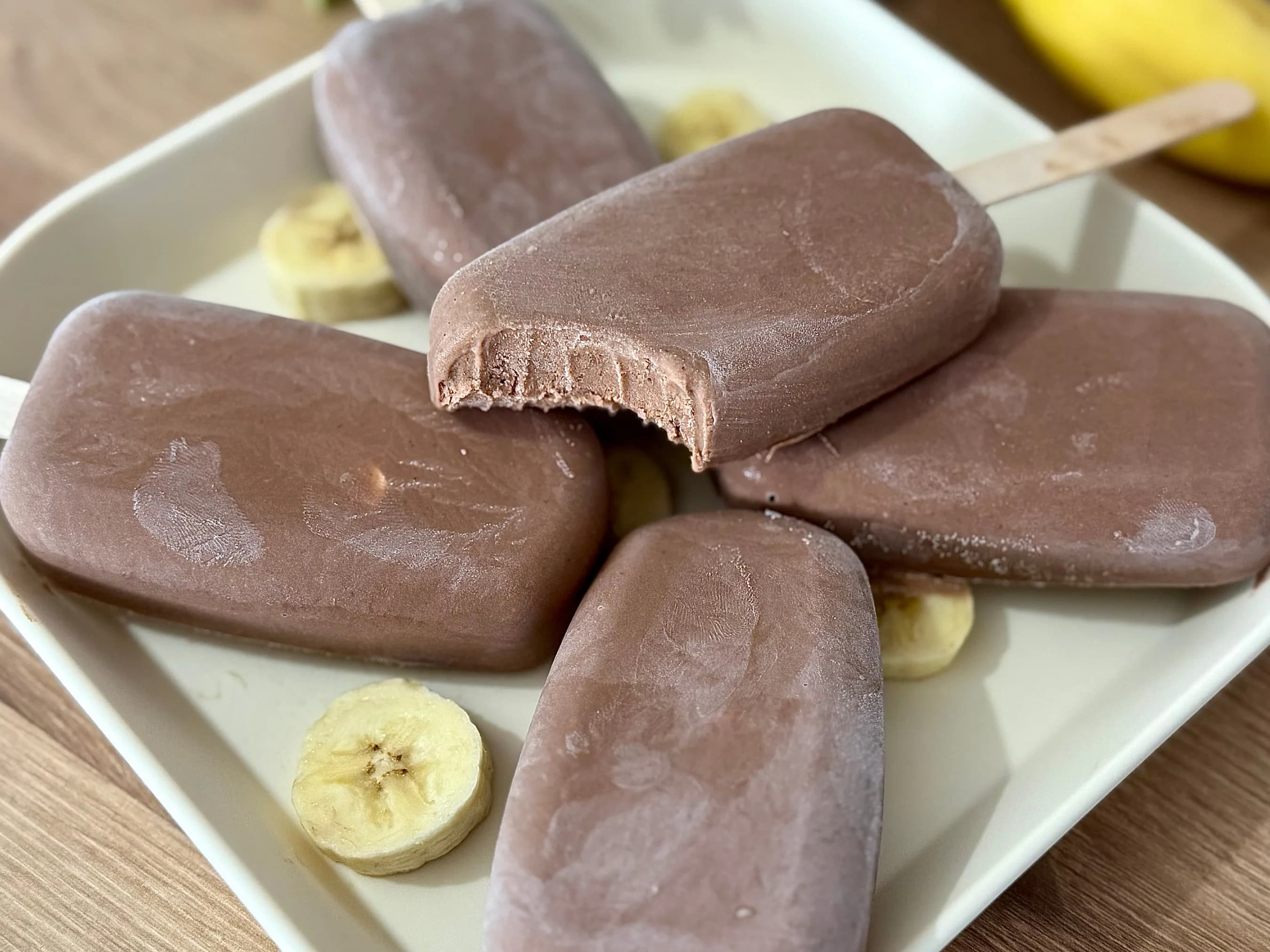 "Creamy chocolate fudge popsicles on a white plate with banana slices, one popsicle has a bite taken out of it. Ideal for a refreshing summer dessert or healthy snack."