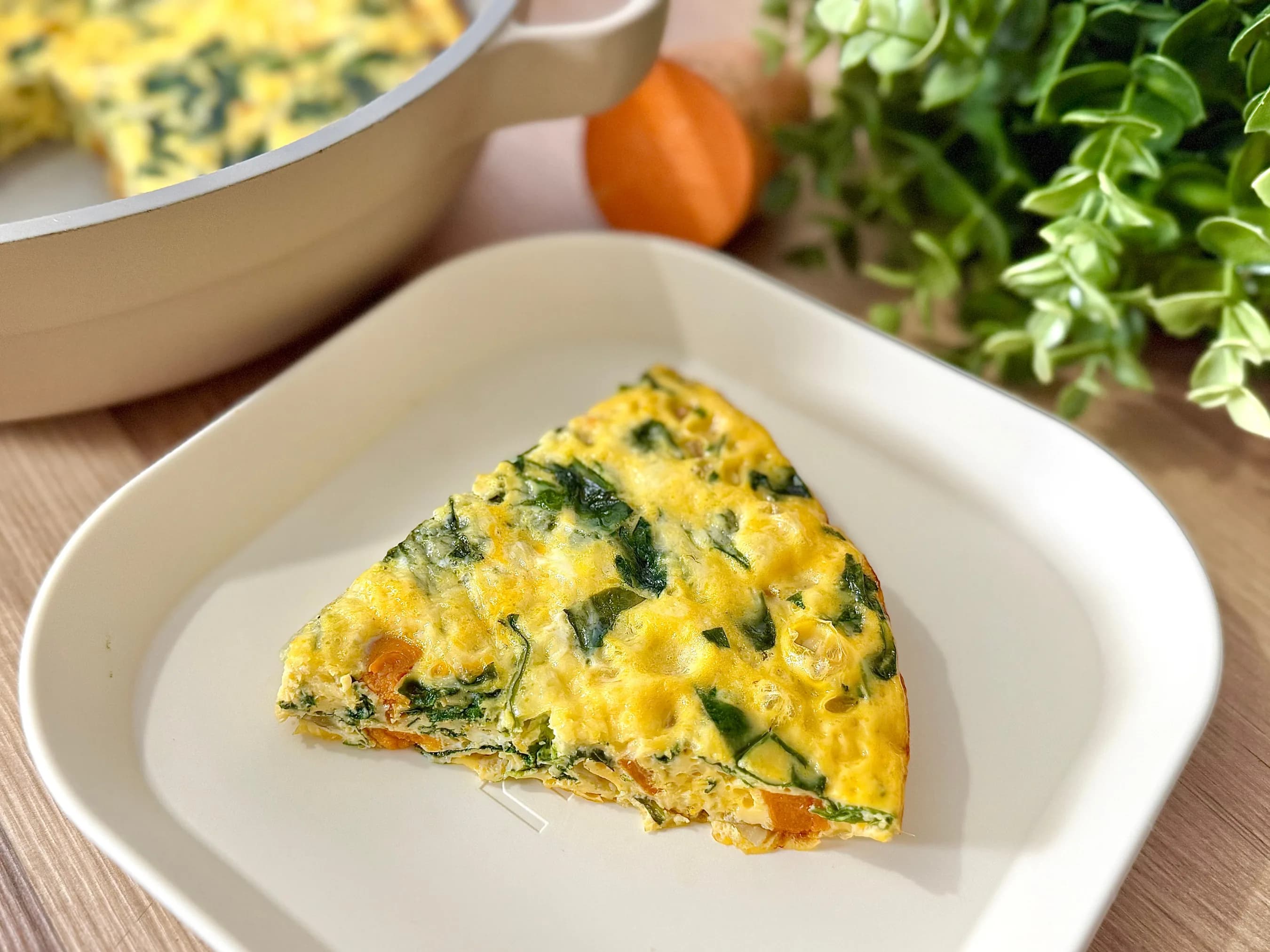 A vibrant slice of potato frittata sits on a white rectangular plate, showcasing a golden-yellow color with flecks of green spinach and diced orange sweet potato. In the background, a large beige pan holds the rest of the frittata, with fresh sweet potato, herbs, and leafy greens visible.