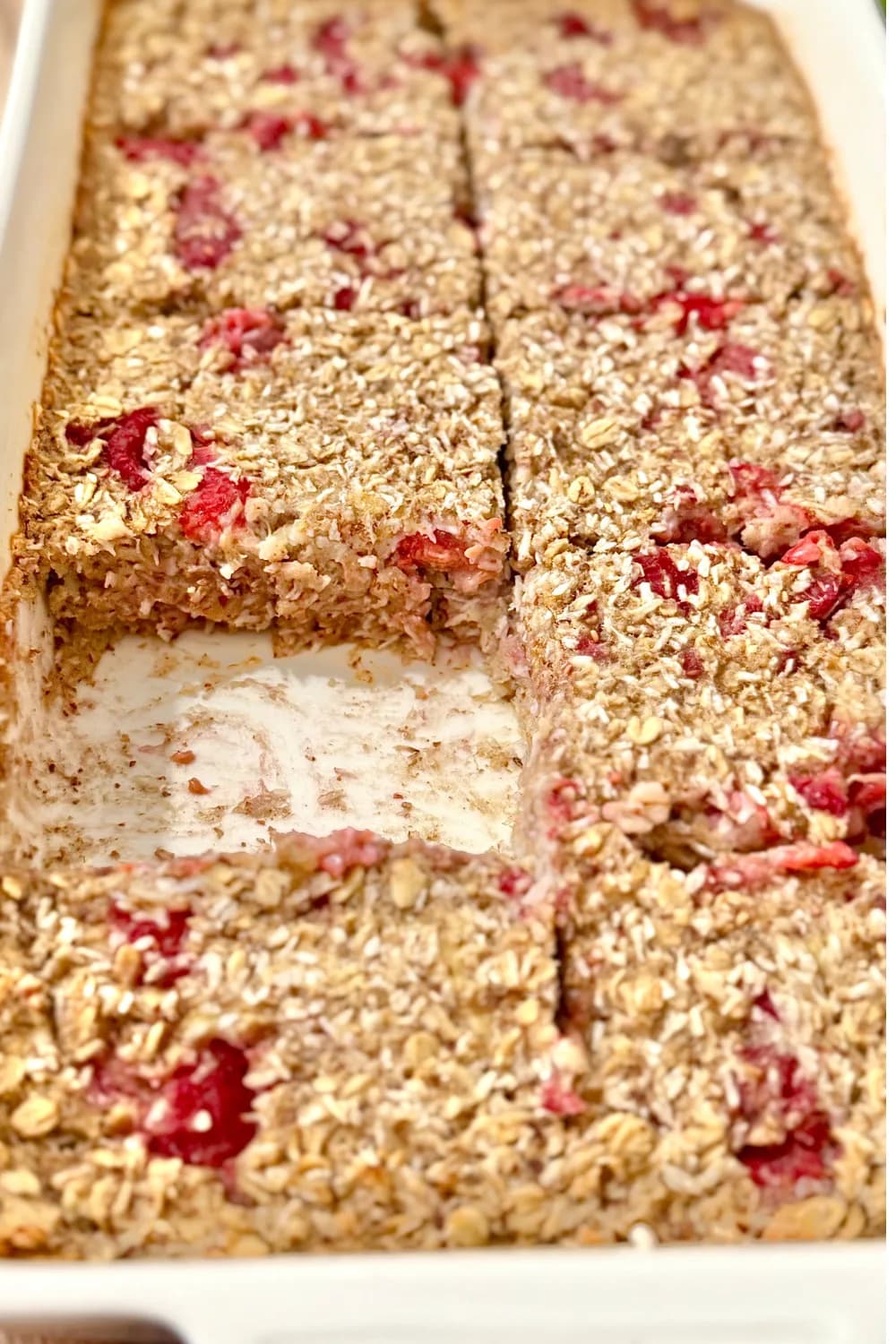 Baked coconut and raspberry oat bars in a white dish, with one square removed to reveal the chewy texture and visible fruit pieces.