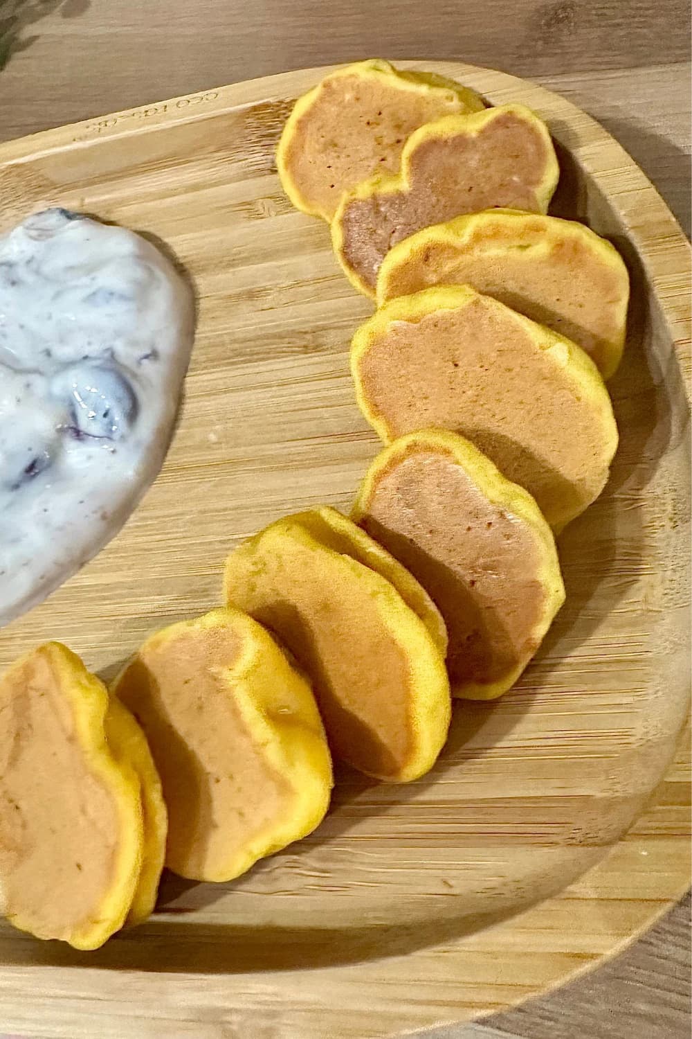 Overhead view of a wooden serving tray with a row of golden mini pumpkin pancakes and a dollop of white, berry-swirled yogurt.