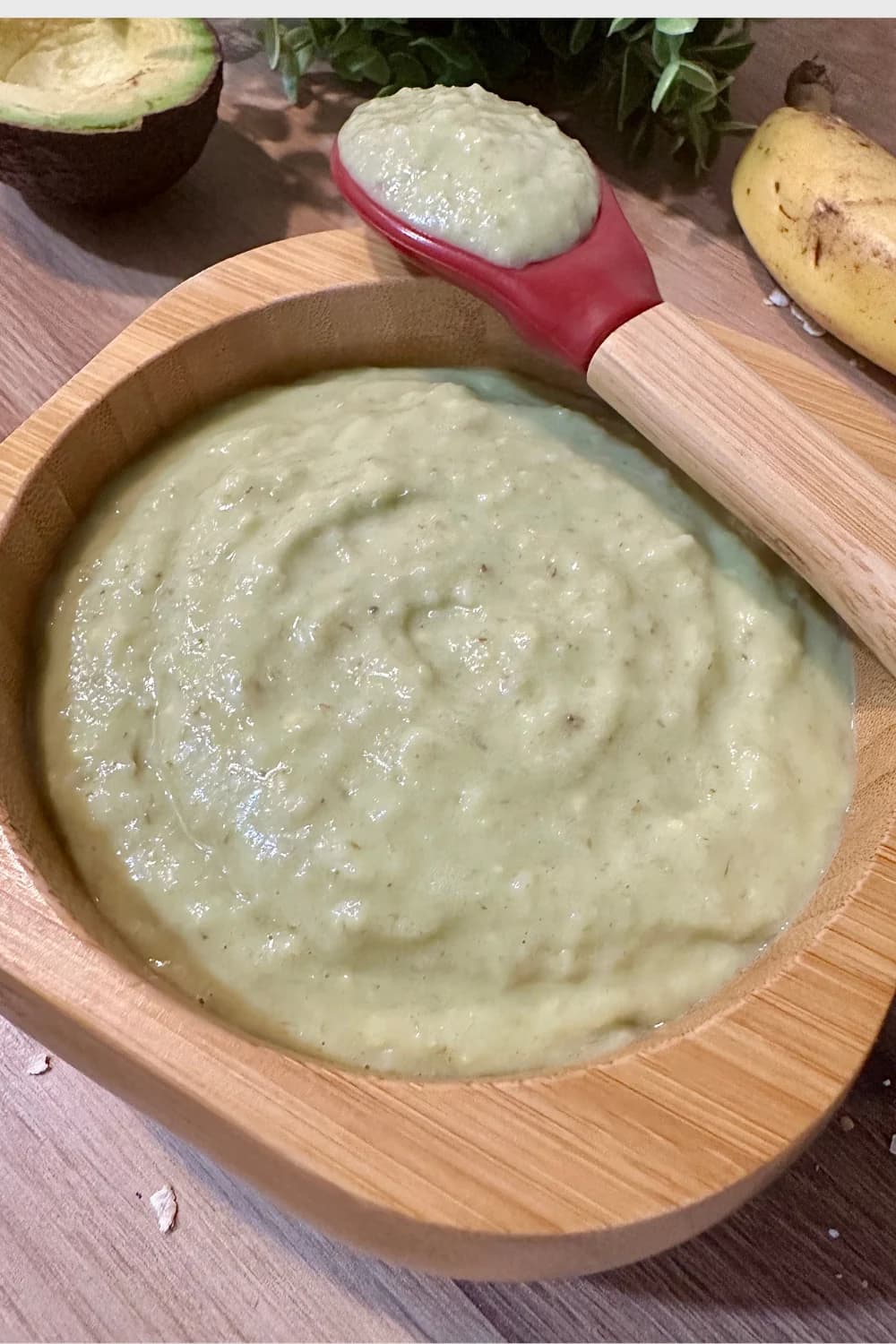Close-up of creamy avocado, banana, and oatmeal baby food puree in a natural bamboo suction bowl, with a red silicone baby spoon resting on the side. A peeled avocado half and a ripe banana are visible in the background, highlighting fresh ingredients for healthy infant and toddler meals.