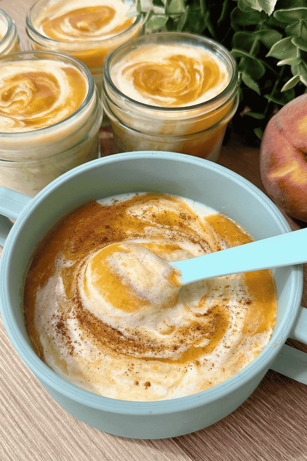 A glass jar and a bowl filled with creamy homemade peach yoghurt, topped with fresh peach slices, with a spoon beside it and ripe peaches in the background, creating a fresh and wholesome presentation.