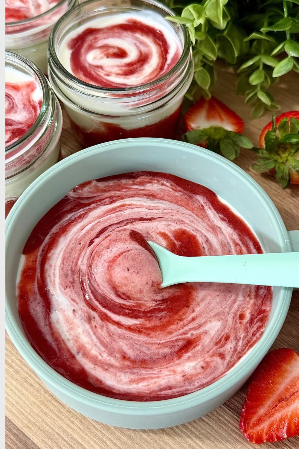 "Overhead close-up of a light blue bowl filled with vibrant strawberry yogurt, featuring a beautiful red and white swirl pattern, with a mint green spoon in the center. Blurry background shows two glass jars of layered strawberry yogurt parfaits and fresh sliced strawberries. Ideal for a healthy breakfast or snack."