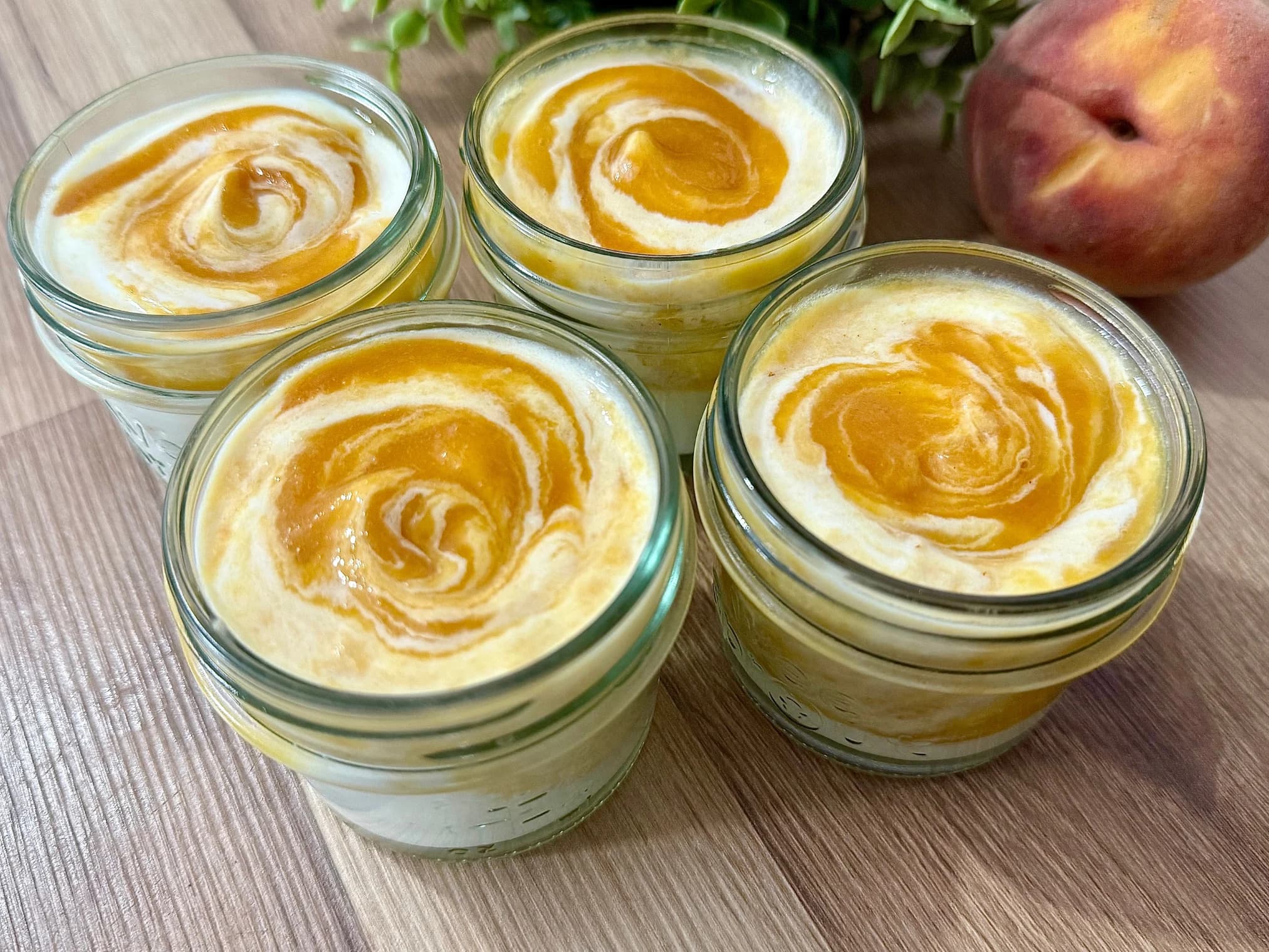 A glass jar filled with creamy homemade peach yoghurt, topped with fresh peach slices, with a spoon beside it and ripe peaches in the background, creating a fresh and wholesome presentation.