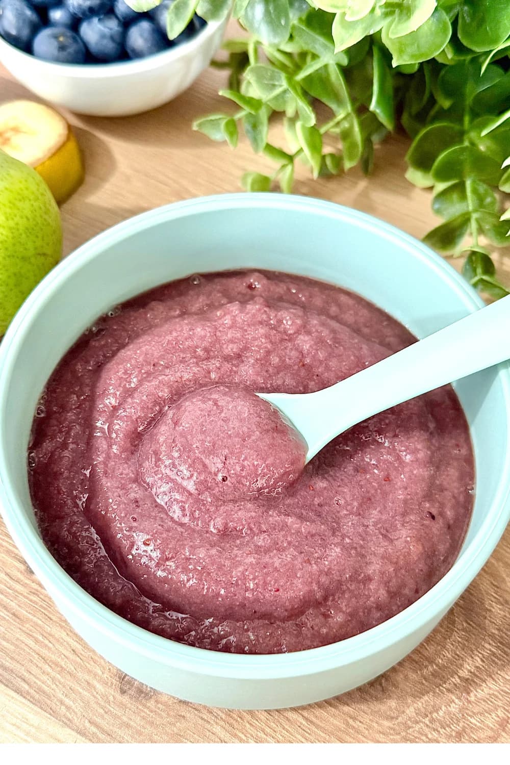 vibrant close-up shot of a light blue bowl filled with Berry Pear Puree. A matching spoon is in the bowl, showing the smooth, thick texture of the purple baby food.