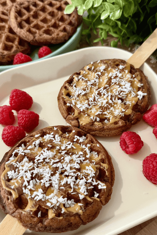 Coconut chocolate waffle pops topped with nut butter and shredded coconut, served on a plate with fresh raspberries.