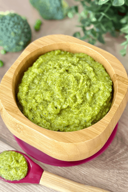 Bowl of green Broccoli & Pea Curry Purée with a bamboo spoon resting on top, placed on a wooden surface with peas and broccoli in the background.