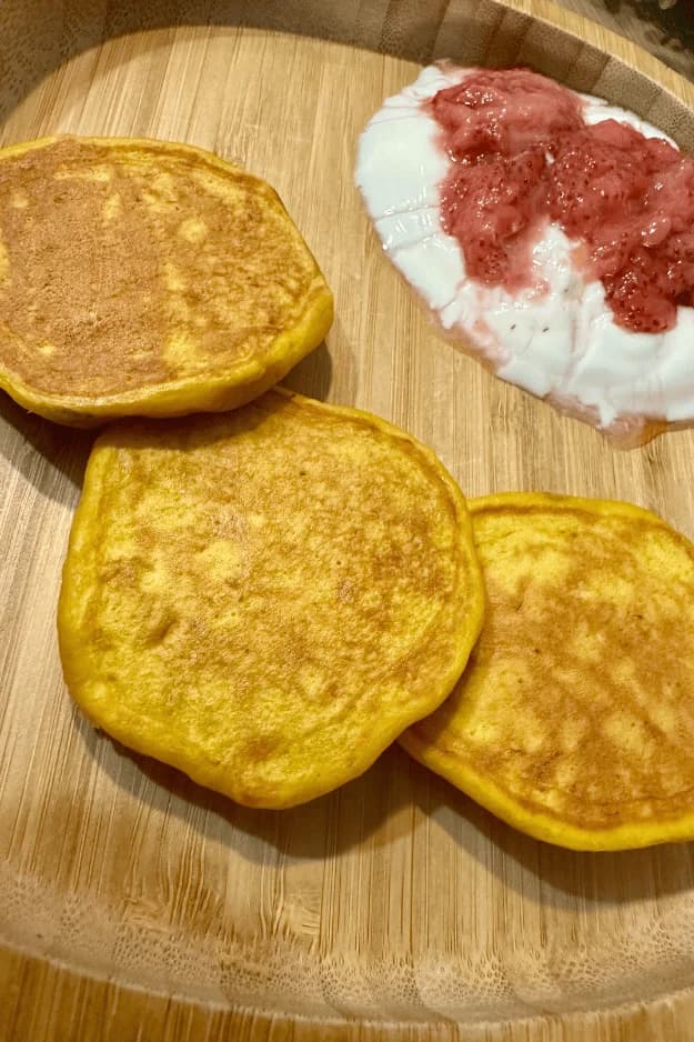 A wooden plate with three fluffy golden pancakes, served with a side of creamy yogurt topped with fresh strawberry sauce. The pancakes have a soft, well-cooked surface, and the yogurt adds a contrast of texture. Designed by Mindsmaking.