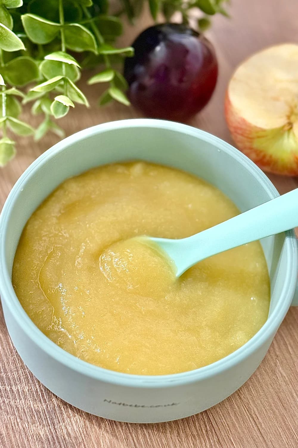 A light blue baby bowl filled with a smooth, yellow apple and plum puree. A matching light blue spoon is resting in the bowl. In the blurred background, there is a whole dark purple plum, a half-cut apple showing its core, and some green leafy plants. The bowl is on a light wooden surface.