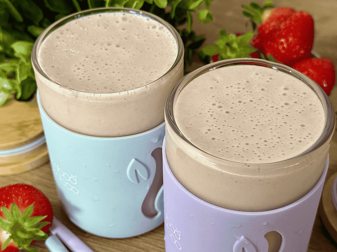 Two glasses of creamy strawberry banana smoothie in pastel-colored sleeves, surrounded by fresh strawberries and green leaves on a wooden surface.