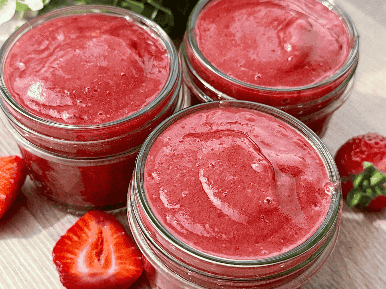 Three small glass jars filled with a smooth, vibrant homemade pink strawberry jam, surrounded by fresh strawberries on a light wooden surface.