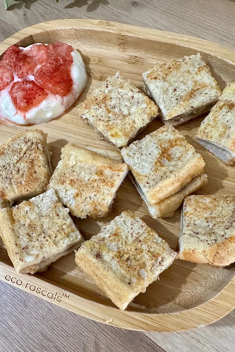 Overhead view of a wooden "eco rascals" plate with neatly arranged squares of banana French toast and a side of yogurt topped with mashed strawberries, ideal for baby-led weaning.Banana-Stuffed French Toast Recipe, suitable for babies 6 months and older. Ingredients displayed include milk, egg, whole grain bread, banana, olive oil, and a prepared dish of banana-stuffed French toast pieces with a side of yogurt and strawberries.