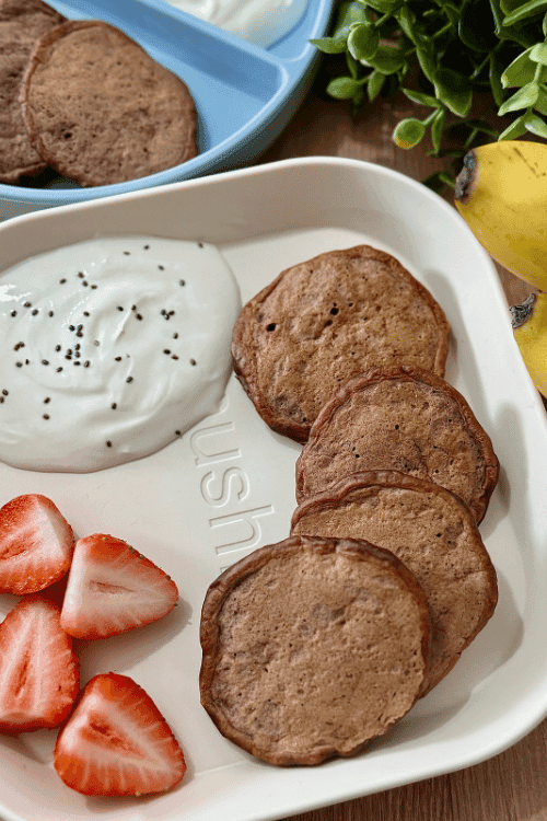 Plate of easy chocolate banana pancakes served with fresh strawberry slices and a dollop of yogurt topped with chia seeds.