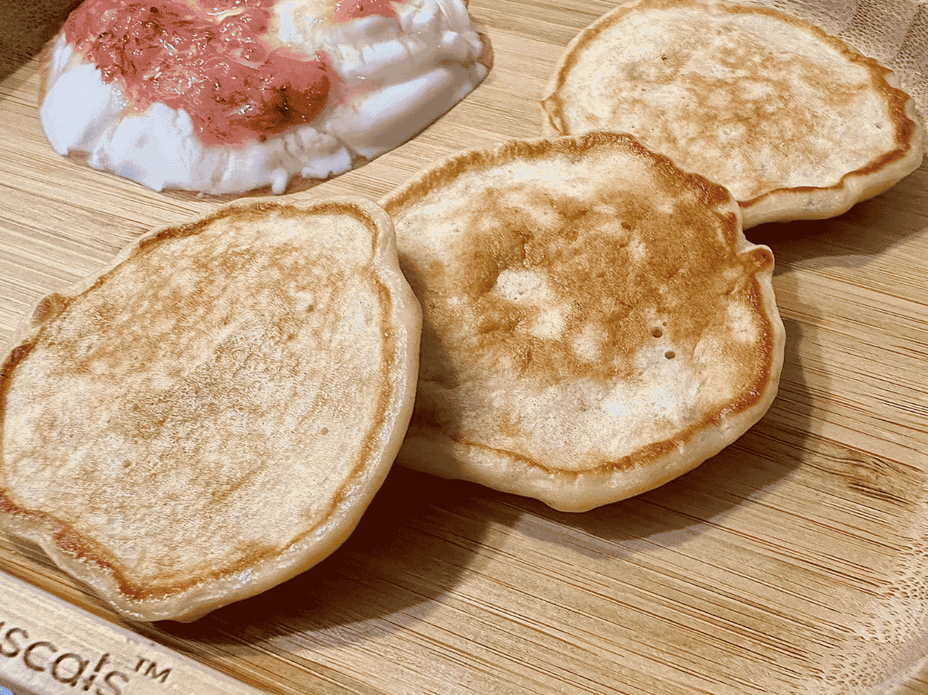 A stack of fluffy golden-brown banana pancakes made with Greek yoghurt, topped with banana slices and a drizzle of syrup, served on a white plate for a wholesome breakfast or snack.