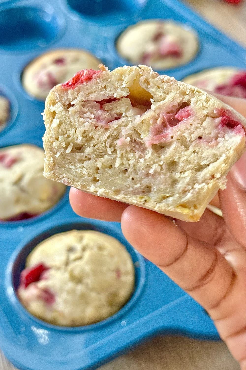 A person's hand holds a half of a freshly baked raspberry muffin, showing the soft, textured interior with swirls of bright pink raspberry.